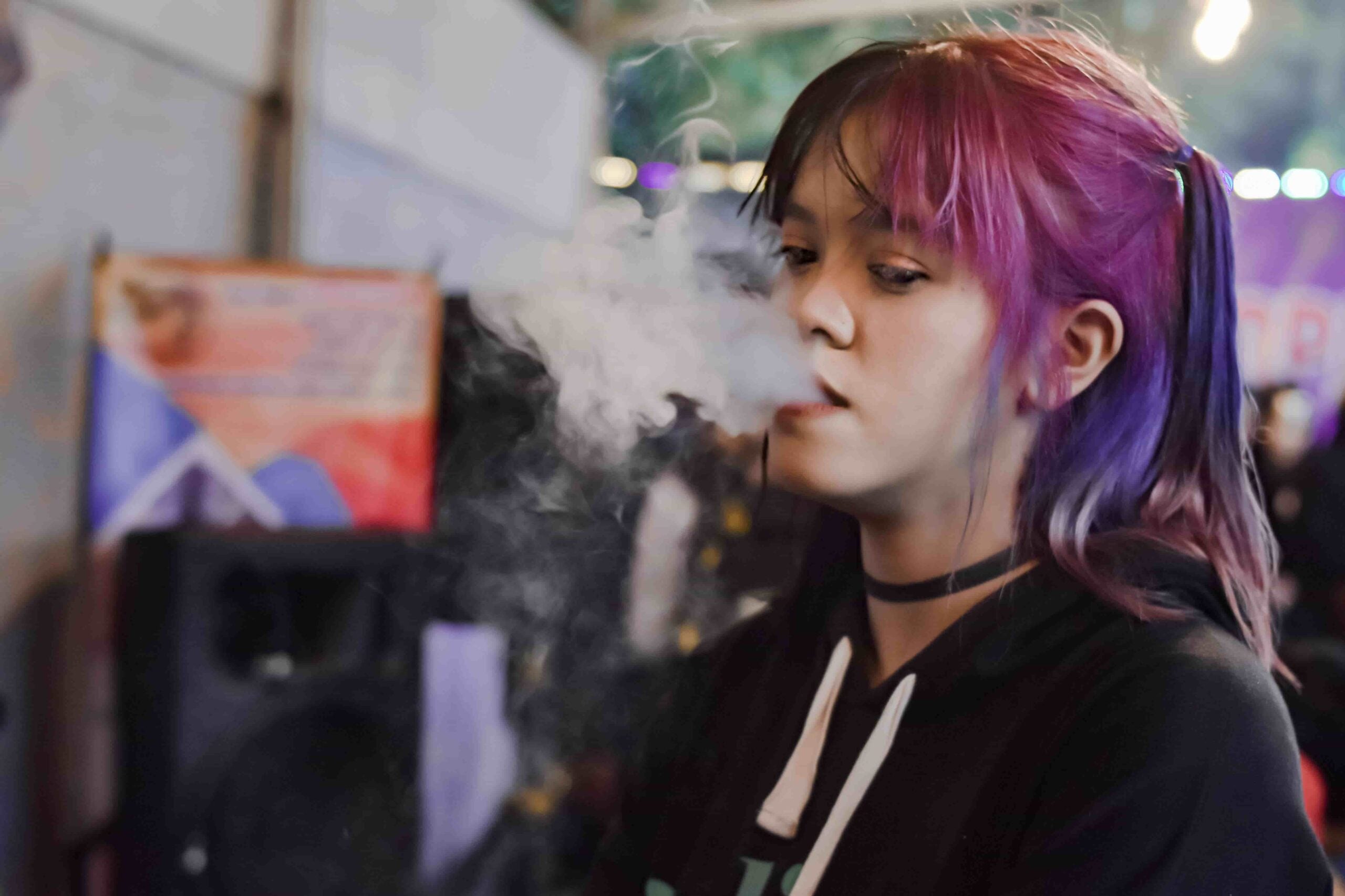Young woman with vibrant purple and pink hair vaping Clicky indoors, exhaling a thick cloud of vapor in a dimly lit environment with colorful lights in the background.