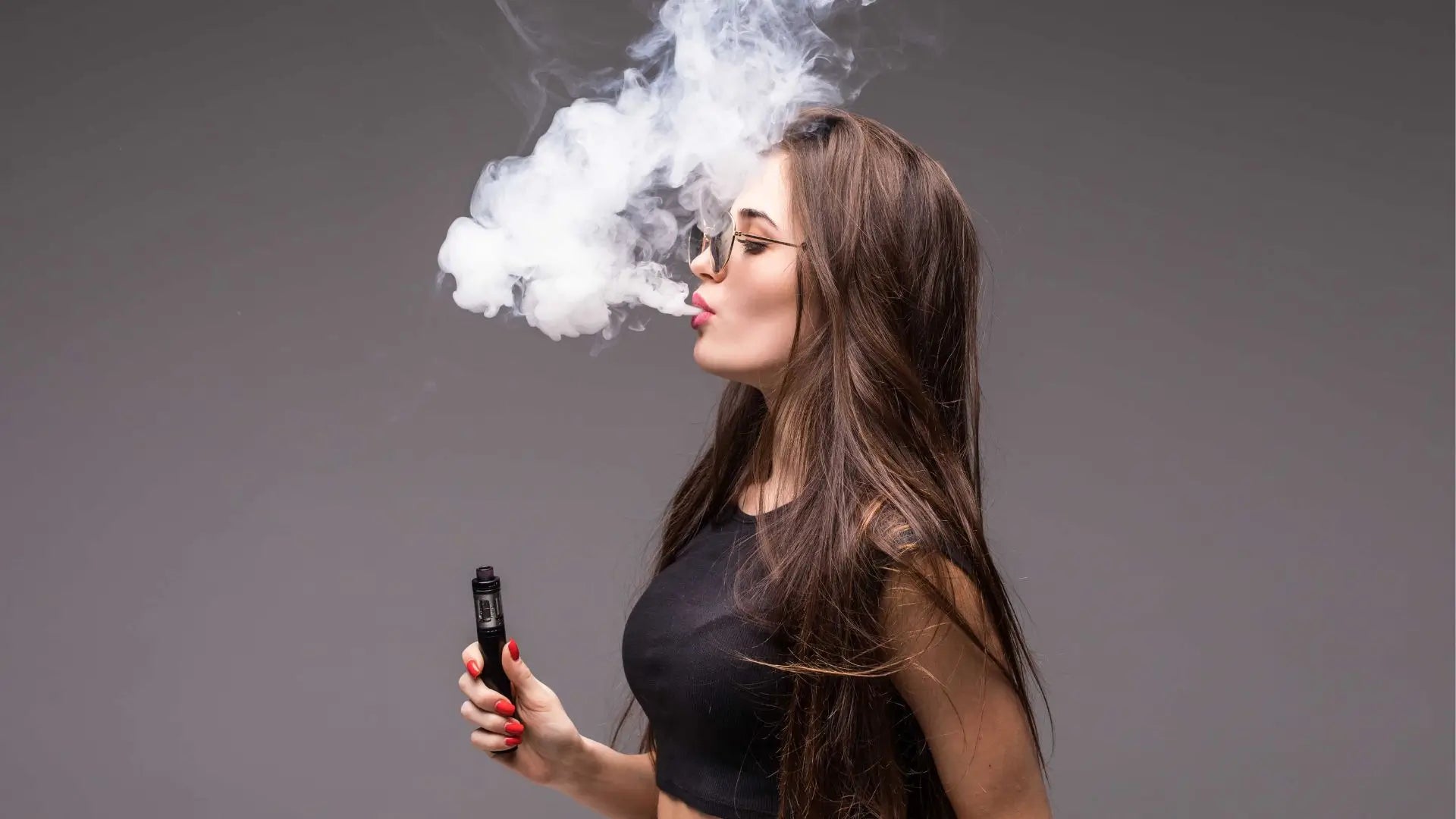Young woman in sunglasses and black clothes vaping and blowing smoke of clicky vape pod on gray wall background